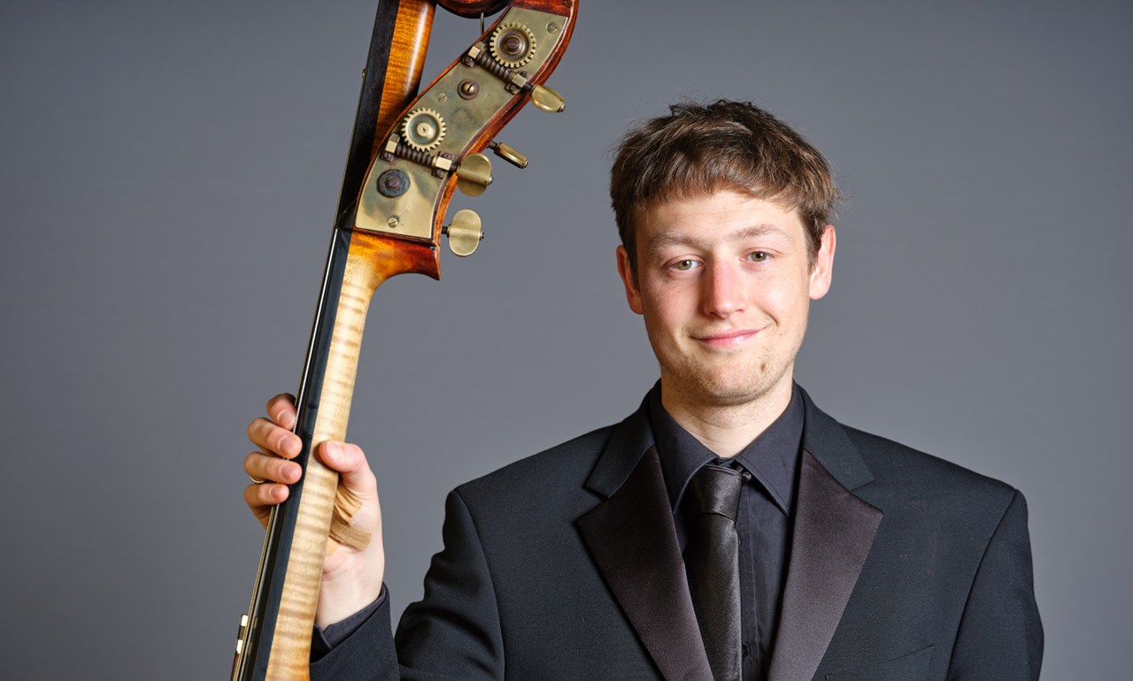 dark haired double bassist holding his instrument in a dark suit